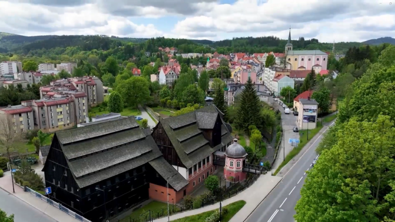 Widok z góry na malowniczą panoramę miasta z dominującym na pierwszym planie zabytkowym Młynem Papierniczym. Budynek młyna ma unikalną, wielopoziomową konstrukcję z ciemnego drewna i spadziste dachy kryte gontem. Obok znajduje się charakterystyczny różowy pawilon z kopułą. W tle widać kolorowe kamienice, wieżę kościoła oraz gęsto zalesione wzgórza pod błękitnym, lekko zachmurzonym niebem. Całość przecina asfaltowa droga z wyraźnym oznakowaniem poziomym.