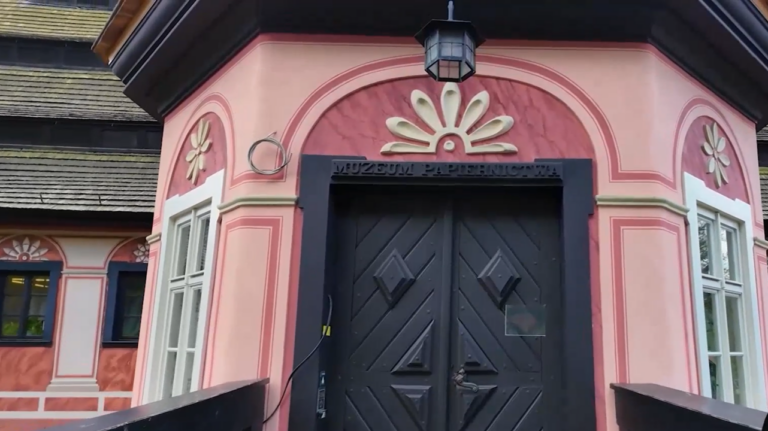 Zbliżenie na ozdobne wejście do Muzeum Papiernictwa. Frontowa ściana jest pomalowana na różowo i ozdobiona klasycznymi sztukateriami oraz białymi detalami w kształcie kwiatów/rozet. Nad ciężkimi, ciemnobrązowymi drewnianymi drzwiami widnieje czarny napis „MUZEUM PAPIERNICTWA”. Po bokach znajdują się wąskie, białe okna, a nad wejściem wisi staromodna, czarna latarnia. W lewym górnym rogu widoczny jest fragment dachu z drewnianego gontu.