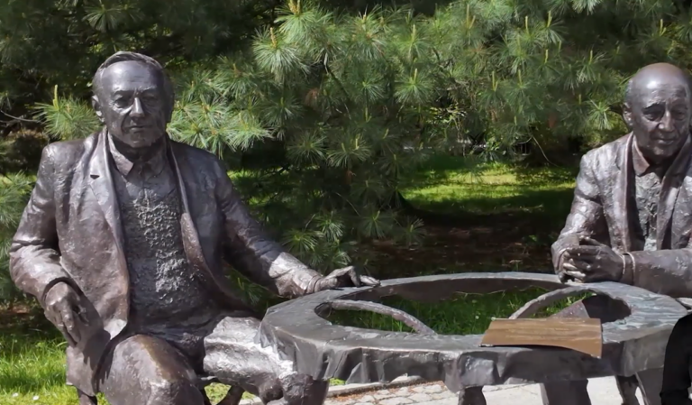 Rzeźba z brązu przedstawiająca dwóch starszych mężczyzn siedzących przy okrągłym stole w parku. Postacie są ubrane w garnitury i siedzą naprzeciwko siebie w swobodnych pozach. Stół ma nieregularny, artystyczny kształt, a na jego blacie leży tabliczka z napisem. W tle widać gęste, zielone iglaki i nasłoneczniony trawnik.