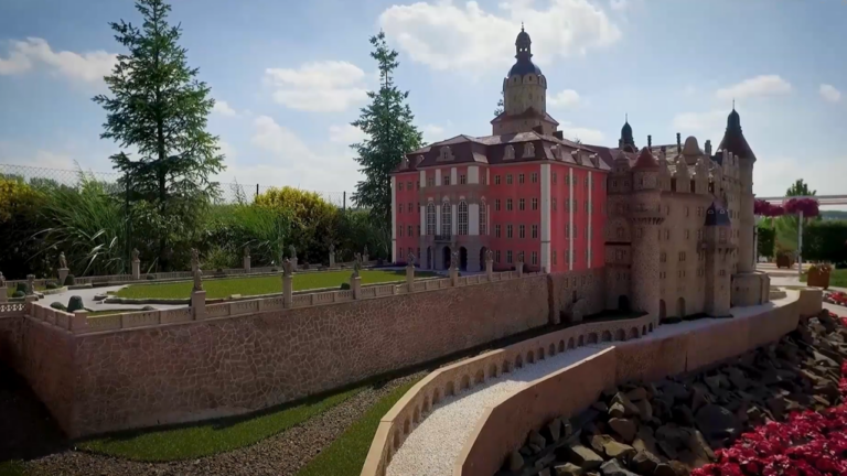 Zdjęcie przedstawia szczegółową miniaturową makietę Zamku Książ, znajdującą się w parku miniatur. Makieta ukazuje charakterystyczną różową fasadę głównego budynku z licznymi oknami oraz przylegające do niego beżowe mury z wieżyczkami i basztami. Przed zamkiem rozciągają się zielone tarasy ogrodowe otoczone niskim murkiem z małymi rzeźbami. W tle widoczne są żywe drzewa i krzewy, które kontrastują z wielkością modelu, a po prawej stronie fragment rabaty z czerwonymi kwiatami. Całość sfotografowana w pełnym słońcu pod jasnym niebem.