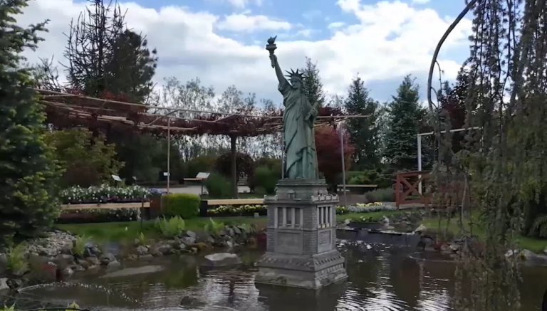 Zdjęcie przedstawia miniaturową replikę Statuy Wolności ustawioną na środku niewielkiego stawu w parku miniatur. Posąg ma charakterystyczny zielonkawy kolor patyny i stoi na szarym, ozdobnym cokole zanurzonym w wodzie. W tle widoczna jest bogata roślinność ogrodowa: iglaki, krzewy oraz drewniana konstrukcja pergoli. Woda w stawie jest spokojna, z delikatnymi kręgami na powierzchni. Całość kompozycji dopełnia jasne, lekko zachmurzone niebo.