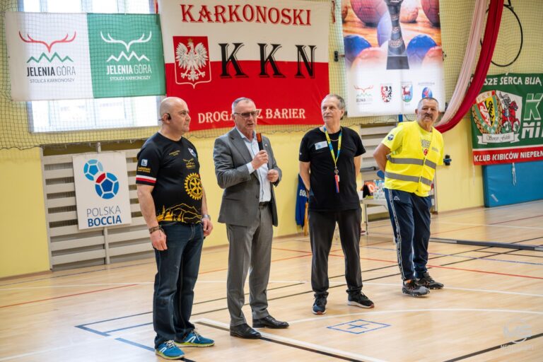 Zdjęcie grupowe czterech mężczyzn stojących na parkiecie sali gimnastycznej podczas wydarzenia sportowego. W centrum starszy mężczyzna w szarym garniturze przemawia do mikrofonu. Po jego lewej stronie stoi mężczyzna w czarnej koszulce z logo "KARKONOSKI KKN" i logiem Rotary. Po prawej stronie mówcy stoją dwaj kolejni mężczyźni: jeden w czarnej koszulce z akredytacją na smyczy, a drugi w odblaskowej, żółtej kamizelce. W tle wiszą transparenty, w tym duży czerwono-biały z napisem "KARKONOSKI KKN" i orłem, oraz zielono-białe flagi "JELENIA GÓRA STOLICA KARKONOSZY". Widoczny jest również baner z napisem "POLSKA BOCCIA".