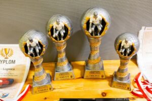 Fotografia wykonana z góry, przedstawiająca cztery pamiątkowe statuetki ustawione na drewnianym stole obok dyplomów. Trofea mają kształt kulistej siatki (przypominającej globus), z której wyłania się srebrzysta sylwetka zawodnika rzucającego kulą. Podstawa każdego trofeum jest srebrno-złota, ze spiralnym, teksturowanym wzorem i prostokątną tabliczką. Tekst na tabliczkach wskazuje miejsce: III MIEJSCE, II MIEJSCE, I MIEJSCE i IV MIEJSCE w turnieju boccia, z dopiskiem "JELENIOGÓRSKI TURNIEJ BOCCI". Obok trofeów leży dyplom z wizerunkiem medalu i wieńca laurowego.