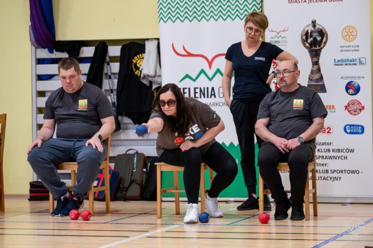 Ujęcie z boku, na którym zawodniczka z niepełnosprawnością, siedząca na małym, drewnianym krześle, wykonuje precyzyjny rzut niebieską kulą. Jest skupiona, w okularach przeciwsłonecznych, ubrana w brązową koszulkę. Obok niej siedzą dwaj mężczyźni w szarych koszulkach z logo (jeden po lewej, drugi po prawej), a za nimi stoi kobieta w czarnej koszulce i okularach, obserwująca rzut. Na parkiecie leżą już rzucone czerwone kule. W tle widać baner z logiem "JELENIA GÓRA KARKONOSZE" i logiem Rotary Club.