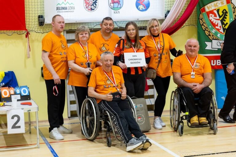 Zdjęcie grupowe sześciu osób ubranych w jednolite, jaskrawopomarańczowe koszulki z logo turnieju na piersi. Drużyna pozuje na tle ściany z banerami sponsorów i klubów (m.in. JELENIA GÓRA, WKS ŚLĄSK). Trzy osoby stoją, dwie siedzą na wózkach inwalidzkich, a jeden mężczyzna klęczy pośrodku. Mężczyzna klęczący trzyma białą kartkę z napisem "KKN CHROBREGO GŁOGÓW". Większość członków drużyny ma na szyi medale na czerwono-białych tasiemkach. Po lewej stronie widać stolik sędziowski z tablicą wyników i numerem "2".
