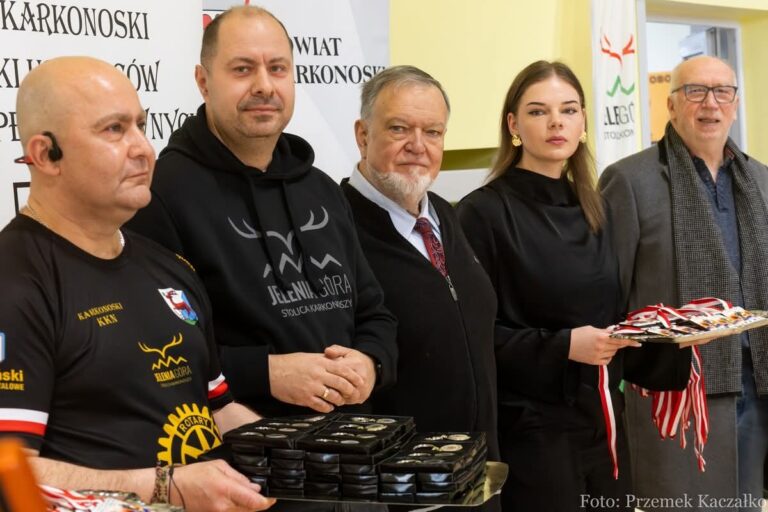 Zbliżenie na moment wręczania nagród. Pięć osób stoi w rzędzie, zwróconych w bok. Na pierwszym planie mężczyzna w czarnej koszulce "KARKONOSKI KKN" z logiem Rotary trzyma czarną tackę z medalami lub przypinkami. Obok niego stoi wyższy mężczyzna w czarnej bluzie z kapturem z napisem "JELENIA GÓRA STOLICA KARKONOSZY". Dalej stoją starszy mężczyzna z brodą i młoda kobieta w czarnej sukience, która trzyma srebrną tacę pełną medali na czerwono-białych wstążkach. Po prawej stronie widać starszego mężczyznę w szarym płaszczu. Tło stanowią banery.