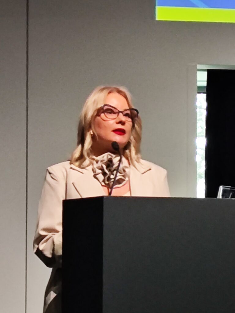 Portret prelegentki o blond włosach i w okularach, przemawiającej zza ciemnej mównicy. Kobieta ma na sobie jasną marynarkę i ozdobny kwiatowy element pod szyją.