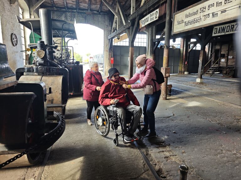 Zdjęcie przedstawia grupę trzech osób wewnątrz dużego, ceglanego hangaru lub parowozowni. Na pierwszym planie mężczyzna na wózku inwalidzkim, któremu towarzyszy kobieta w różowej kurtce, poprawiająca mu kołnierz lub smycz na szyi. Obok nich stoi starsza kobieta w czerwonej kurtce. Po lewej stronie widać fragmenty zabytkowych maszyn kolejowych, w tym walec drogowy, a w tle tablice informacyjne z nazwami stacji (m.in. Jaworzyna Śląska).