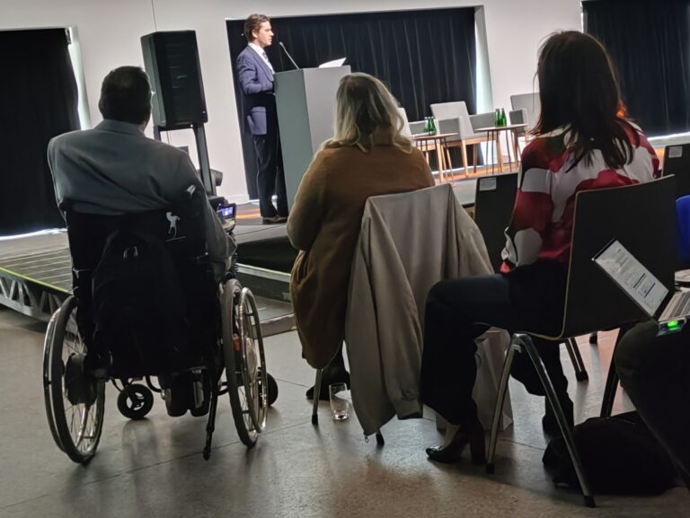 Widok zza pleców uczestników na prelegenta w garniturze stojącego przy mównicy na niskim podeście. Po lewej stronie widać osobę na wózku inwalidzkim, a po prawej siedzące kobiety, z których jedna ma na sobie biało-czerwoną bluzkę w geometryczne wzory.