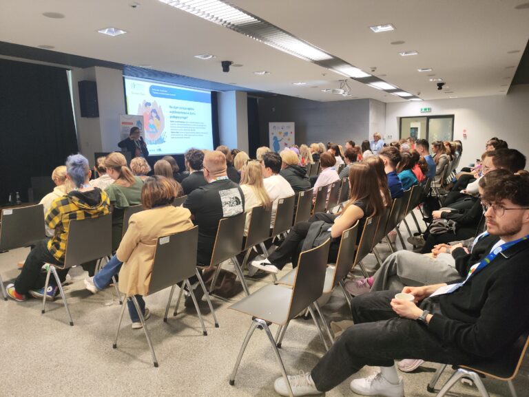 Szerokie ujęcie sali konferencyjnej. Uczestnicy siedzą w rzędach, zwróceni w stronę ekranu, na którym wyświetlana jest prezentacja z grafiką przedstawiającą rodzinę. Na pierwszym planie po prawej widać młodego mężczyznę w okularach trzymającego papierowy kubek.