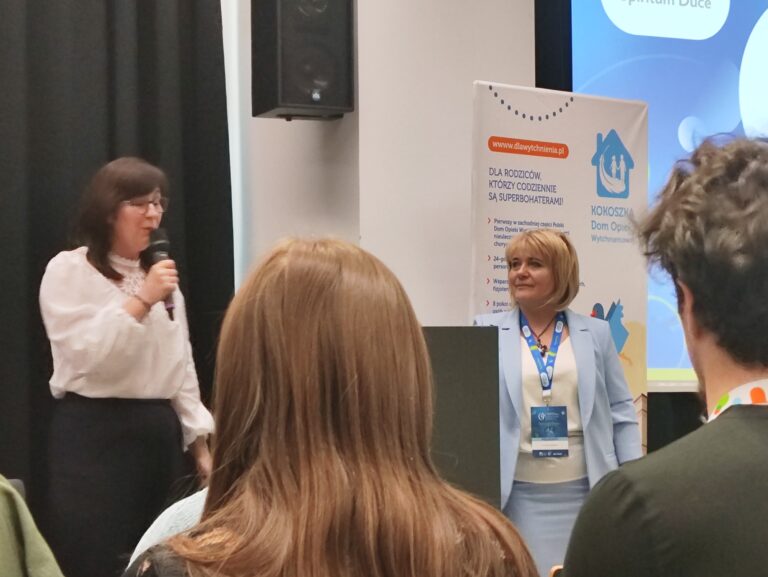 Dwie kobiety na froncie sali. Jedna z nich mówi do mikrofonu, ubrana w białą, koronkową bluzkę. Druga stoi obok, ubrana w błękitną marynarkę z identyfikatorem na szyi. W tle widoczny jest roll-up „Kokoszka Dom Opieki Wytchnieniowej”.