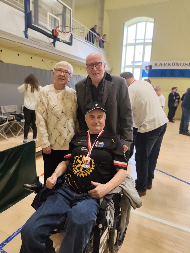 Portret trzech osób stojących w hali sportowej. Po lewej stronie starsza kobieta w kremowym swetrze i okularach z czerwoną oprawką, po prawej starszy mężczyzna w szarym garniturze i okularach, a pośrodku mężczyzna w średnim wieku na wózku inwalidzkim w czarnej koszulce i czapce z daszkiem. Kobieta i mężczyzna w garniturze patrzą w obiektyw, mężczyzna na wózku patrzy lekko w dół.