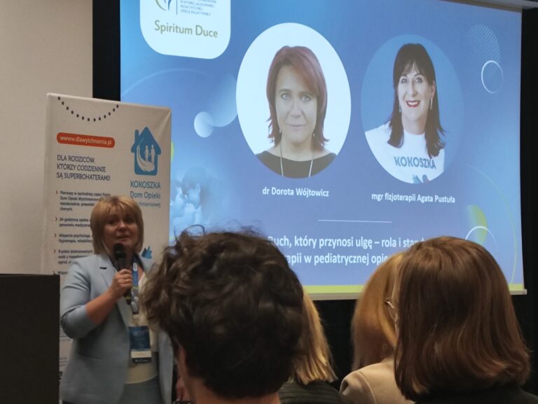 Prelegentka w błękitnej marynarce przemawia do mikrofonu na tle slajdu prezentacji. Na ekranie widoczne są dwa portrety kobiet: dr Doroty Wójtowicz oraz mgr Agaty Pustuły, a pod nimi tytuł wystąpienia dotyczący roli fizjoterapii.