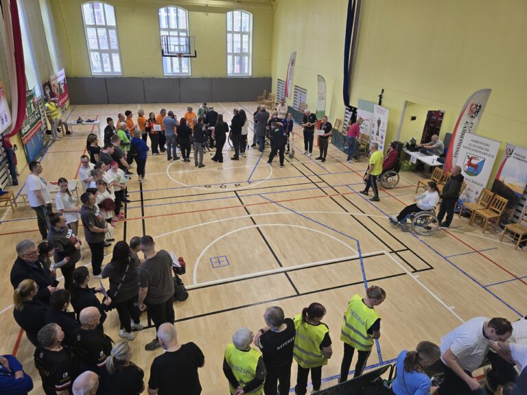 Ujęcie z góry na halę sportową, gdzie na parkiecie odbywają się zawody w boccia. Liczne grupy osób w sportowych strojach (czarnych, pomarańczowych, białych) stoją wokół wyznaczonych boisk. Widać sędziów w odblaskowych kamizelkach i kilka osób na wózkach inwalidzkich. Na ścianie wiszą banery.