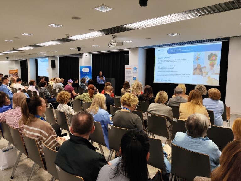 Szerokie ujęcie sali konferencyjnej wypełnionej słuchaczami. Na froncie prelegentka stoi obok dużego ekranu wyświetlającego prezentację z napisem „Dzień w Piórku” oraz zdjęciami dzieci. W tle widoczne są logotypy organizatorów.