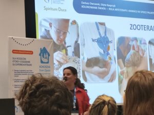Prelegentka w czerwonej koszuli przemawia na tle slajdu zatytułowanego „Zooterapia”. Na ekranie wyświetlone są zdjęcia przedstawiające kontakt pacjentów ze zwierzętami, m.in. z świnką morską i dużym ślimakiem.