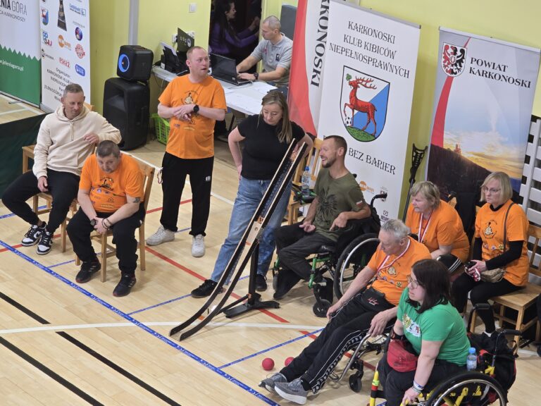 Ujęcie z bliska na grupę osób siedzących i stojących na parkiecie podczas zawodów. W centrum widać rurę startową do boccia. Obok rury siedzi kobieta na wózku inwalidzkim, a za nią stoi mężczyzna na wózku. Widać również osoby stojące w pomarańczowych i czarnych koszulkach oraz banery w tle.