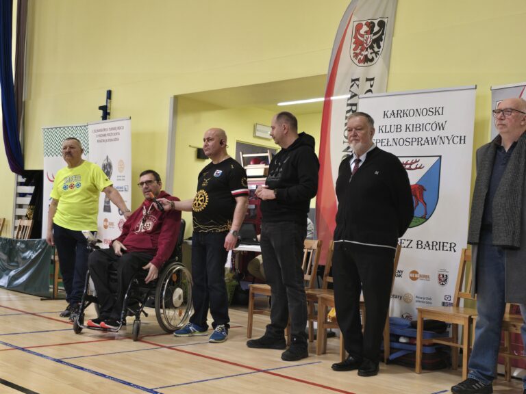 Zdjęcie z poziomu parkietu na trzech zawodników grających w boccia. Dwaj starsi mężczyźni siedzą na krzesłach i rzucają kulami. Trzeci mężczyzna, również na krześle, przygotowuje się do rzutu. Na parkiecie leży kilka czerwonych i niebieskich kul. W tle banery z logotypami.