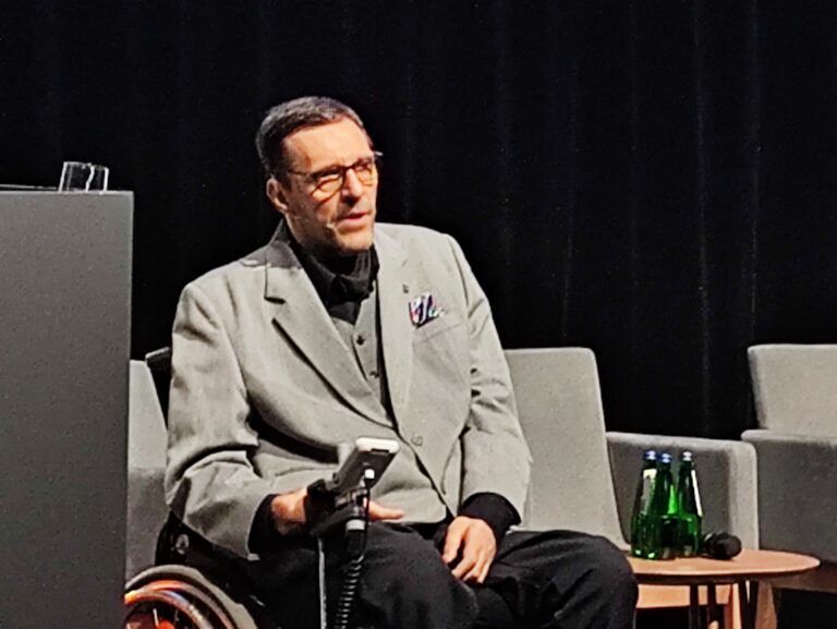 Mężczyzna w okularach i szarym garniturze siedzący na wózku inwalidzkim podczas panelu dyskusyjnego. W ręku trzyma mikrofon, a obok na stoliku stoją butelki z wodą.