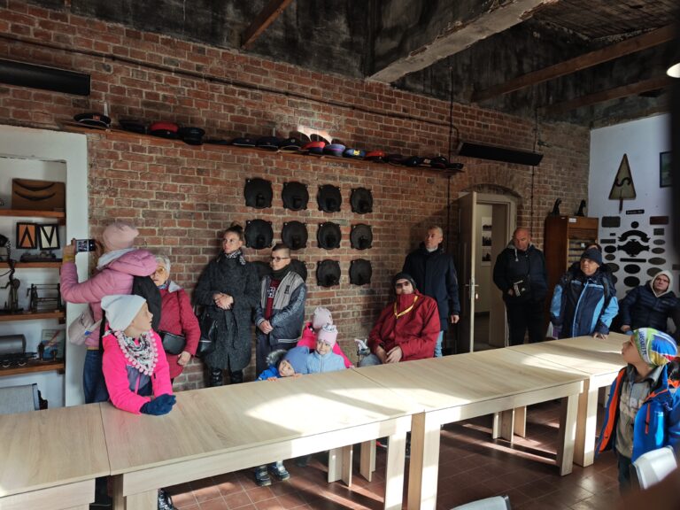 Grupowe zdjęcie dorosłych i dzieci zgromadzonych w pomieszczeniu o surowych, ceglanych ścianach. Grupa stoi za długimi, jasnymi stołami, patrząc w jednym kierunku, prawdopodobnie słuchając przewodnika. Na ścianie w tle wiszą liczne eksponaty kolejowe: czapki mundurowe na górnej półce oraz rzędy ciemnych, metalowych elementów (prawdopodobnie tabliczek lub części maszyn). Wnętrze ma charakter edukacyjny, muzealny.