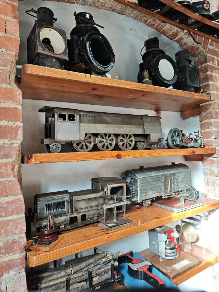 Zbliżenie na drewniane regały wbudowane w ceglaną wnękę, na których wyeksponowano zabytkowe przedmioty kolejowe. Na najwyższej półce stoją duże, czarne lampy parowozowe. Poniżej znajdują się metalowe modele pociągów i wagonów o surowym, technicznym wyglądzie. Na najniższych poziomach widać mniejsze elementy mechaniczne, przekroje urządzeń oraz miniaturowe bale drewna na wagoniku.