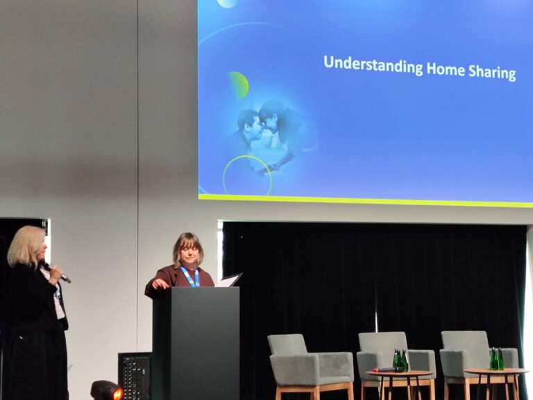 Dwie kobiety na scenie obok ekranu z napisem „Understanding Home Sharing”. Jedna z nich stoi przy mównicy, druga obok z mikrofonem. W tle widać puste fotele konferencyjne.
