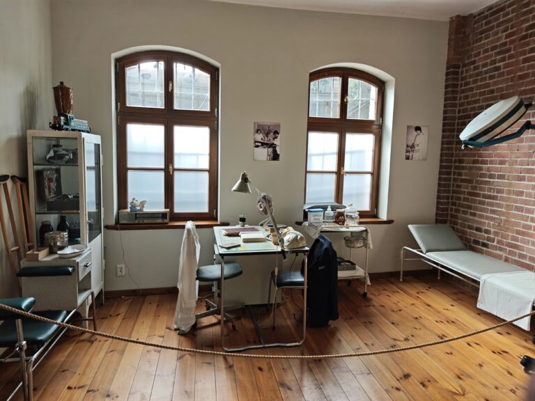 Zdjęcie przedstawia muzealną rekonstrukcję dawnego gabinetu lekarskiego lub kolejowej przychodni. W jasnym pomieszczeniu o drewnianej podłodze znajdują się: metalowe biurko z lampką i starym telefonem, biała szafka medyczna z przeszklonymi drzwiami, leżanka lekarska pod ceglaną ścianą oraz kule ortopedyczne oparte o ścianę. Nad leżanką wisi duża, okrągła lampa operacyjna. Pomieszczenie jest oddzielone od zwiedzających grubym sznurem.