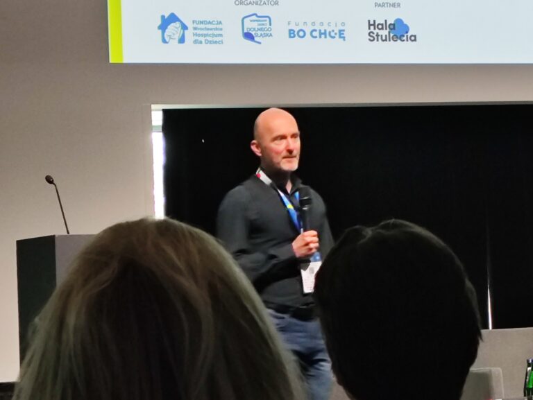 Zbliżenie na uśmiechniętego prelegenta w ciemnej koszuli, trzymającego mikrofon. W górnej części kadru widoczne fragmenty logotypów fundacji i partnerów wydarzenia.