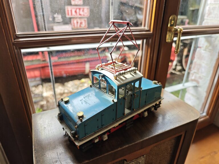 Zbliżenie na precyzyjny model niebieskiej lokomotywy elektrycznej z czerwonym pantografem na dachu. Model stoi na drewnianej obudowie starego radia. W tle, przez szybę okienną, widać fragmenty prawdziwego parowozu o oznaczeniu TKt48-173, co podkreśla skalę i charakter muzeum.