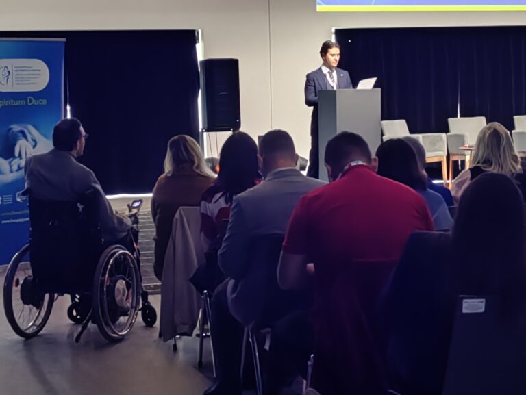 Widok z perspektywy uczestników na mężczyznę w garniturze przemawiającego przy mównicy. W pierwszym planie widać tyły głów słuchaczy oraz osobę na wózku inwalidzkim tyłem do obiektywu.