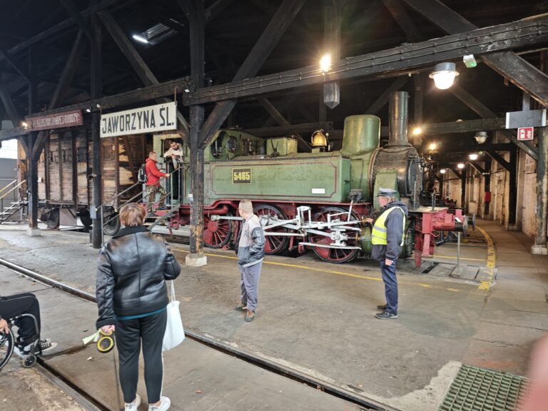 Wnętrze ciemnej, drewnianej parowozowni. Na torze stoi zielono-czarny parowóz o numerze 5465. Przed maszyną stoi pracownik w odblaskowej kamizelce, a obok przechodzą zwiedzający. Nad pociągiem widać białą tablicę z napisem „JAWORZYNA ŚL.”. Konstrukcja dachu jest wsparta na masywnych, czarnych belkach.