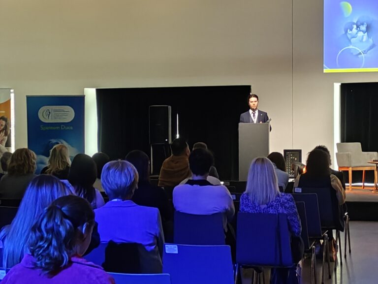 Szerokie ujęcie sali konferencyjnej w niebieskim oświetleniu. Na scenie prelegent przy mównicy, a przed nim rzędy siedzących uczestników wydarzenia.