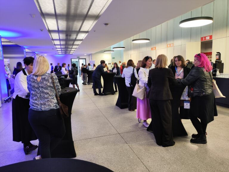 Uczestnicy konferencji podczas przerwy kawowej w nowoczesnym lobby. Ludzie stoją w grupach przy wysokich, czarnych stolikach koktajlowych, rozmawiając i pijąc napoje.