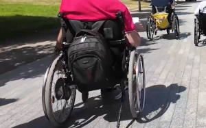 Widok od tyłu na grupę osób poruszających się na wózkach inwalidzkich po jasnej, brukowanej alejce w słoneczny dzień. Na pierwszym planie widać osobę w różowej koszulce z czarnym plecakiem zawieszonym na oparciu wózka. W oddali wzdłuż ścieżki jadą kolejne dwie osoby.