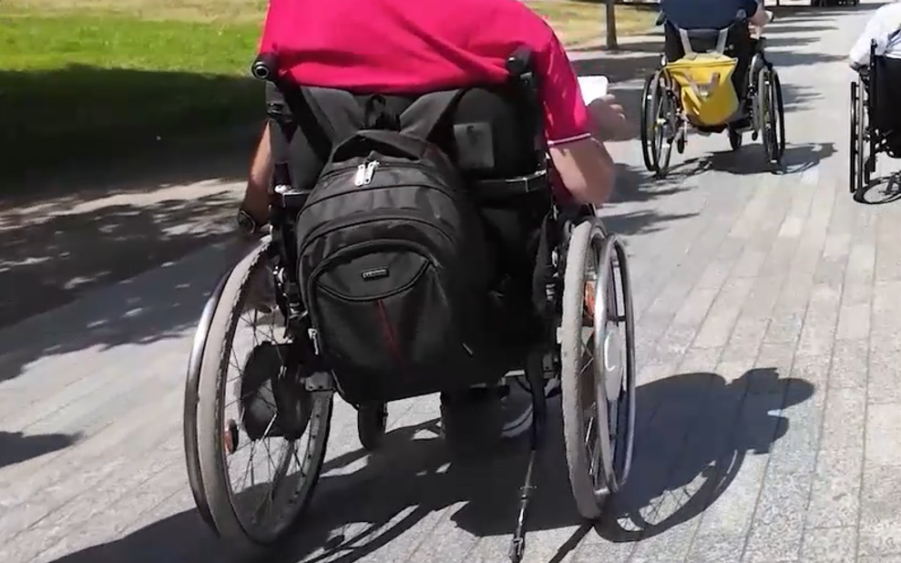 Widok od tyłu na grupę osób poruszających się na wózkach inwalidzkich po jasnej, brukowanej alejce w słoneczny dzień. Na pierwszym planie widać osobę w różowej koszulce z czarnym plecakiem zawieszonym na oparciu wózka. W oddali wzdłuż ścieżki jadą kolejne dwie osoby.