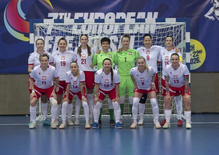 Zdjęcie grupowe żeńskiej reprezentacji Polski w futsalu na tle bramki. Jedenaście zawodniczek w białych koszulkach z czerwonymi detalami i logo orła oraz czerwonych spodenkach. Dwie bramkarki w środku ubrane są w jasnozielone stroje. W tle widać baner „7th European Deaf Futsal Championships”.