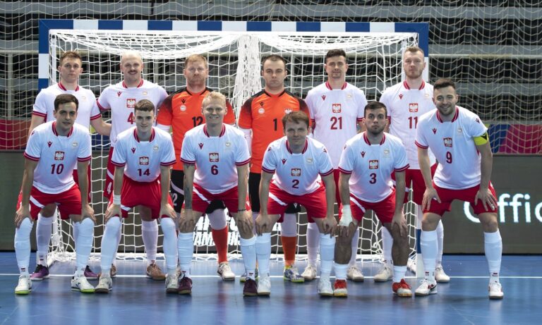 Zdjęcie grupowe męskiej reprezentacji Polski w futsalu. Dwunastu zawodników pozuje przed bramką; siedmiu stoi w tylnym rzędzie, pięciu kuca z przodu. Zawodnicy z pola mają białe koszulki z czerwonymi wykończeniami i czerwone spodenki. Dwaj bramkarze w środku noszą pomarańczowe bluzy.
