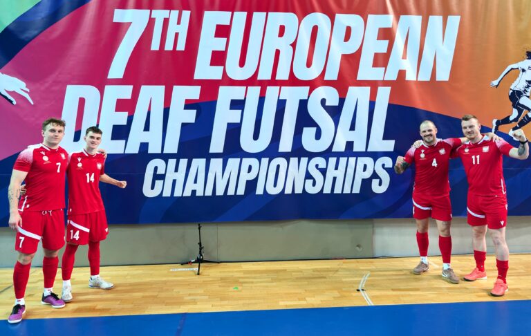 Czterech zawodników męskiej reprezentacji Polski pozuje na hali sportowej przed wielkim banerem „7th European Deaf Futsal Championships”. Zawodnicy są ubrani w czerwone stroje reprezentacyjne i uśmiechają się, wykonując gesty zwycięstwa.