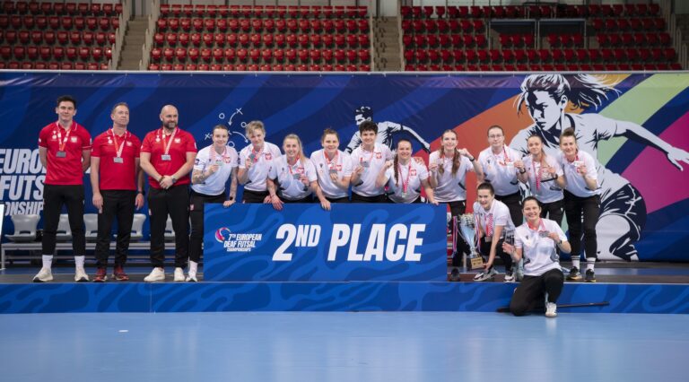 Szerokie ujęcie całej żeńskiej kadry narodowej wraz ze sztabem szkoleniowym na podium. Grupa stoi za niebieską tablicą z napisem „2nd PLACE”. Wszyscy mają na szyjach srebrne medale. W tle widać trybuny hali sportowej oraz grafikę promującą 7. Mistrzostwa Europy Głuchych w Futsalu.