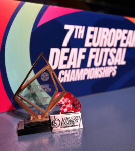 Zbliżenie na trofea sportowe ustawione na niebieskiej nawierzchni. Po lewej stronie znajduje się szklana statuetka „Best Young Player Women”, a obok niej leży srebrny medal „7th European Deaf Futsal Championships” na biało-czerwonej wstążce. W rozmytym tle widoczny jest baner mistrzostw.