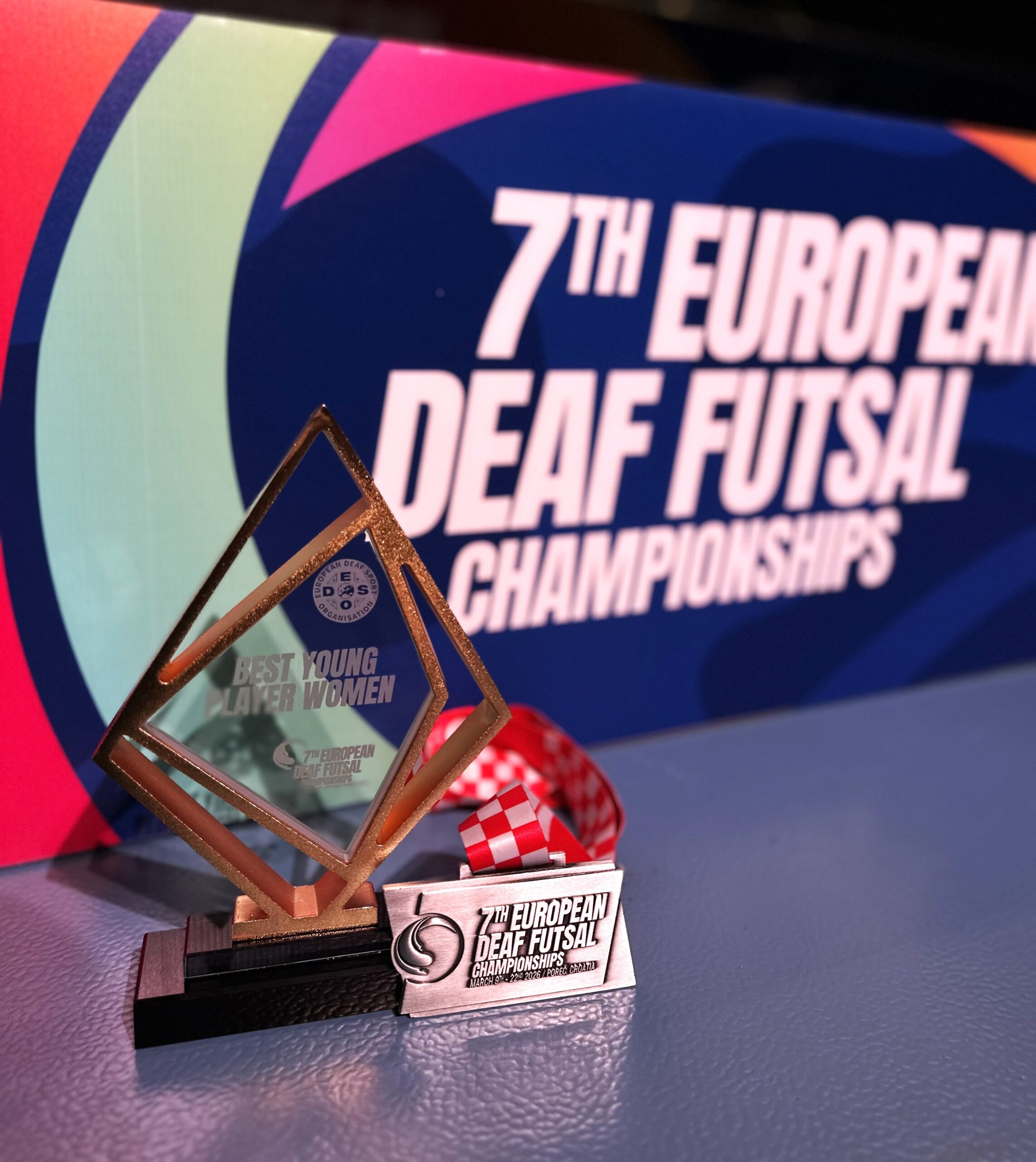 Zbliżenie na trofea sportowe ustawione na niebieskiej nawierzchni. Po lewej stronie znajduje się szklana statuetka „Best Young Player Women”, a obok niej leży srebrny medal „7th European Deaf Futsal Championships” na biało-czerwonej wstążce. W rozmytym tle widoczny jest baner mistrzostw.