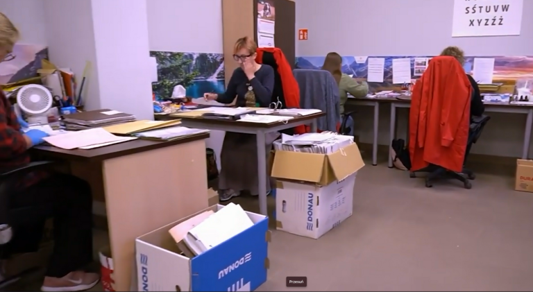Wnętrze biura, w którym kilka osób pracuje przy biurkach nad dokumentacją. Na pierwszym planie widać duże kartonowe pudła marki Donau wypełnione dokumentami. Jedna z kobiet w okularach siedzi przy środkowym biurku i przegląda dokumenty, podczas gdy inne osoby pracują przy sąsiednich stanowiskach. Na ścianie w tle wisi plansza z polskim alfabetem oraz fototapeta przedstawiająca leśny krajobraz.