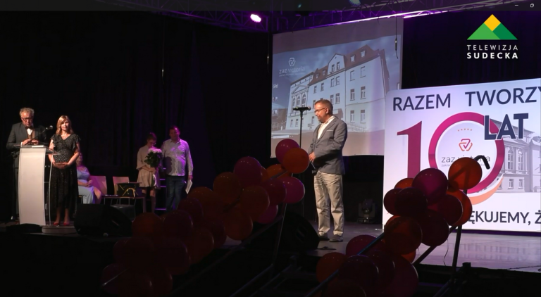 Scena teatru lub sali widowiskowej podczas uroczystej gali z okazji 10-lecia „ZAZ Victoria”. Mężczyzna w jasnych spodniach i szarej marynarce przemawia do mikrofonu na środku sceny. W tle wyświetlany jest slajd z budynkiem instytucji, a po prawej stronie stoi duża ścianka jubileuszowa. Scenę zdobią girlandy z ciemnoczerwonych i pomarańczowych balonów, a po lewej stronie w głębi stoją inni uczestnicy uroczystości.