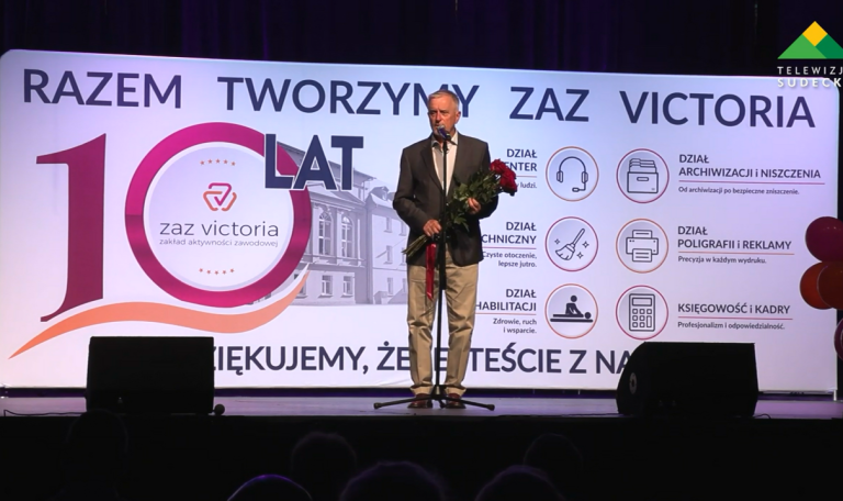 Mężczyzna w garniturze i jasnych spodniach stoi na scenie przed dużą tablicą informacyjną i trzyma w rękach bukiet czerwonych róż. Tablica za nim przedstawia strukturę organizacyjną „ZAZ Victoria” z podziałem na działy: Dział Center, Dział Techniczny, Dział Rehabilitacji, Dział Archiwizacji i Niszczenia, Dział Poligrafii i Reklamy oraz Księgowość i Kadry – każdy zilustrowany odpowiednią ikoną.