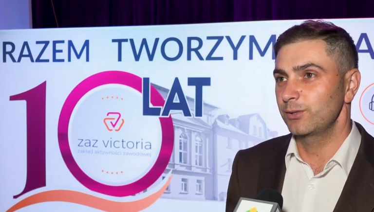 Portretowe zbliżenie na mężczyznę w brązowej marynarce i białej koszuli, który udziela wypowiedzi do mikrofonu (widocznego u dołu kadru). W tle znajduje się biała ścianka z różowym, jubileuszowym logo „10 LAT zaz victoria”.